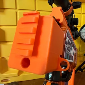 May include: An orange toy gun with black accents and a black clock on a tripod stand. The toy gun has a rectangular design with a series of ridges on top. The clock has a white face with Roman numerals and black hands.