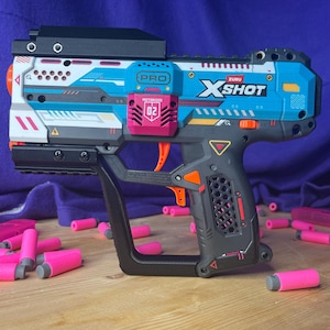 以下が含まれることがあります： 青、白、黒のZuru X-Shot Pro電動おもちゃブラスター。02とラベル付けされたピンクのアクセントパーツが付いています。ブラスターにはオレンジ色のトリガーとテクスチャ加工されたグリップがあります。おもちゃのベースの周りには、小さなピンクのフォームダーツがいくつか散らばっています。