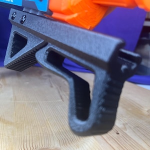 May include: A black angled foregrip attachment for a toy blaster, featuring a textured gripping surface and a hollow triangular design. The accessory is mounted to the underside of a blue and orange toy blaster with two visible black screws.