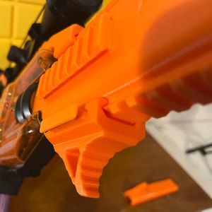 May include: Close-up of an orange and clear toy rifle with a black scope. The rifle has a textured grip and a magazine. A small orange piece is detached and lying on a brown surface.
