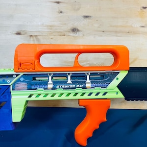 May include: A toy gun with an orange handle and top, and a green and black body. The text "STRYKER 2.0" is visible on the side. The toy gun is set against a wooden background. The design is modern and angular.