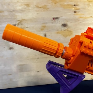May include: An orange and purple toy gun with a cylindrical orange barrel extension. The gun features a textured design and a purple angled grip. The background is a light-colored wooden surface.