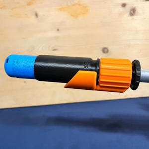 Puede incluir: Primer plano de un accesorio de manguera de jardín. El accesorio tiene una punta azul, un cuerpo negro y un conector naranja. El accesorio está conectado a un poste plateado. El fondo es una superficie de madera.