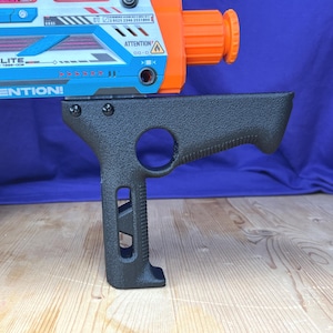 May include: A black textured vertical foregrip attachment mounted to a blue and orange toy blaster. The grip features a circular cutout near the top, a vertical slot in the handle, and two visible screws securing it to the top rail of the blaster.
