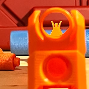 May include: An orange toy gun sight is the focus, featuring a circular aperture and a small, yellow aiming post. The background includes orange and blue cylinders, hinting at a playful or educational context. The scene is set on a light-coloured surface.