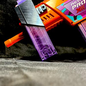 May include: Orange and purple toy gun with the text "NEON PRO XK". The gun has a black magazine and a clear purple magazine with the number "12" on it. The gun has a black and silver top.