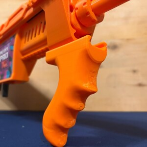 Könnte beinhalten: Nahaufnahme einer orangefarbenen Spielzeugpistole mit strukturiertem Griff. Die Pistole trägt den Text "HEAVY PRO" an der Seite. Der Griff ist mit Fingerrillen versehen. Der Hintergrund ist blau und Holz.