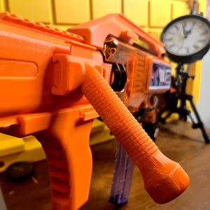 May include: A bright orange toy blaster featuring a textured vertical foregrip, a top rail, and a clear plastic magazine. The blaster has a prominent orange body with visible 3D printed layer lines and a rectangular side panel with graphic details.