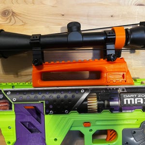May include: A toy gun with a black scope mounted on an orange and green frame. The gun has the text "DART ZONE MAX" on the side. The gun is green, purple, and black.