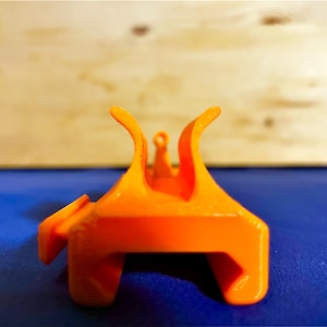 以下が含まれることがあります： ユニークな形状の明るいオレンジ色の3Dプリントオブジェクト。長方形のベースに、上向きに湾曲した要素と上部に小さなループが付いています。オブジェクトは、木製の背景を持つ青い表面に配置されています。