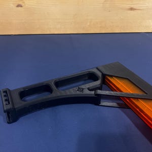 May include: A black and orange object, possibly a component of a larger device, rests on a blue surface. The black part has a geometric design with open sections. The orange section is rectangular and appears to be a magazine.