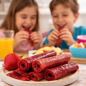 K&ouml;nnte beinhalten: Ein Holzbrettchen zeigt mehrere aufgerollte Fruchtsnacks und einige flache St&uuml;cke. Eine frische Erdbeere liegt neben dem Brettchen. Im Hintergrund essen zwei Kinder die Fruchtsnacks. Ein Glas Orangensaft und eine Obstschale sind ebenfalls zu sehen.