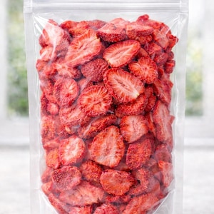 Puede incluir: Una bolsa de plástico transparente llena de rodajas de fresas liofilizadas. Las fresas son de un color rojo vibrante y parecen estar cortadas uniformemente. La bolsa está sellada y lista para consumir.