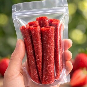K&ouml;nnte beinhalten: Ein klarer Plastikbeutel gef&uuml;llt mit mehreren roten Fruchtlederrollen. Die Rollen sind zylindrisch mit einer strukturierten Oberfl&auml;che und einer leuchtend roten Farbe. Erdbeeren sind im Hintergrund zu sehen, was auf den Geschmack des Fruchtleders hindeutet.