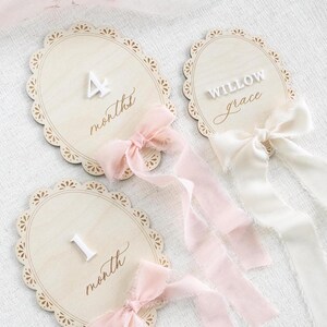 Lace Frame Milestone Marker with Scallop Border. Coquette Bow Detail with block font first name and script middle name or last name or both. Each Lace Frame is adorned with a silk chiffon ribbon in the color light pink or ivory. White acrylic milestone markers with the monthly number interchangeable in acrylic. Matching lace Frame name plate that can be used as a newborn hospital announcement. Milestone marker for girl, bow milestone card for girl, hospital name announcement for girl, wooden