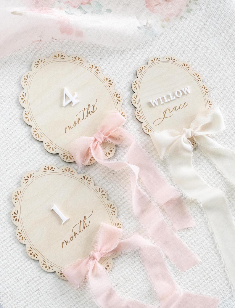 Lace Frame Milestone Marker with Scallop Border. Coquette Bow Detail with block font first name and script middle name or last name or both. Each Lace Frame is adorned with a silk chiffon ribbon in the color light pink or ivory. White acrylic milestone markers with the monthly number interchangeable in acrylic. Matching lace Frame name plate that can be used as a newborn hospital announcement. Milestone marker for girl, bow milestone card for girl, hospital name announcement for girl, wooden