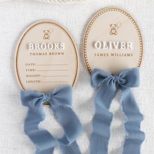 May include: Two wooden oval baby announcement plaques. One reads "BROOKS" and the other "OLIVER" in white block letters. Each has a teddy bear graphic and a light blue ribbon. The "BROOKS" plaque has lines for date, time, weight, and length.