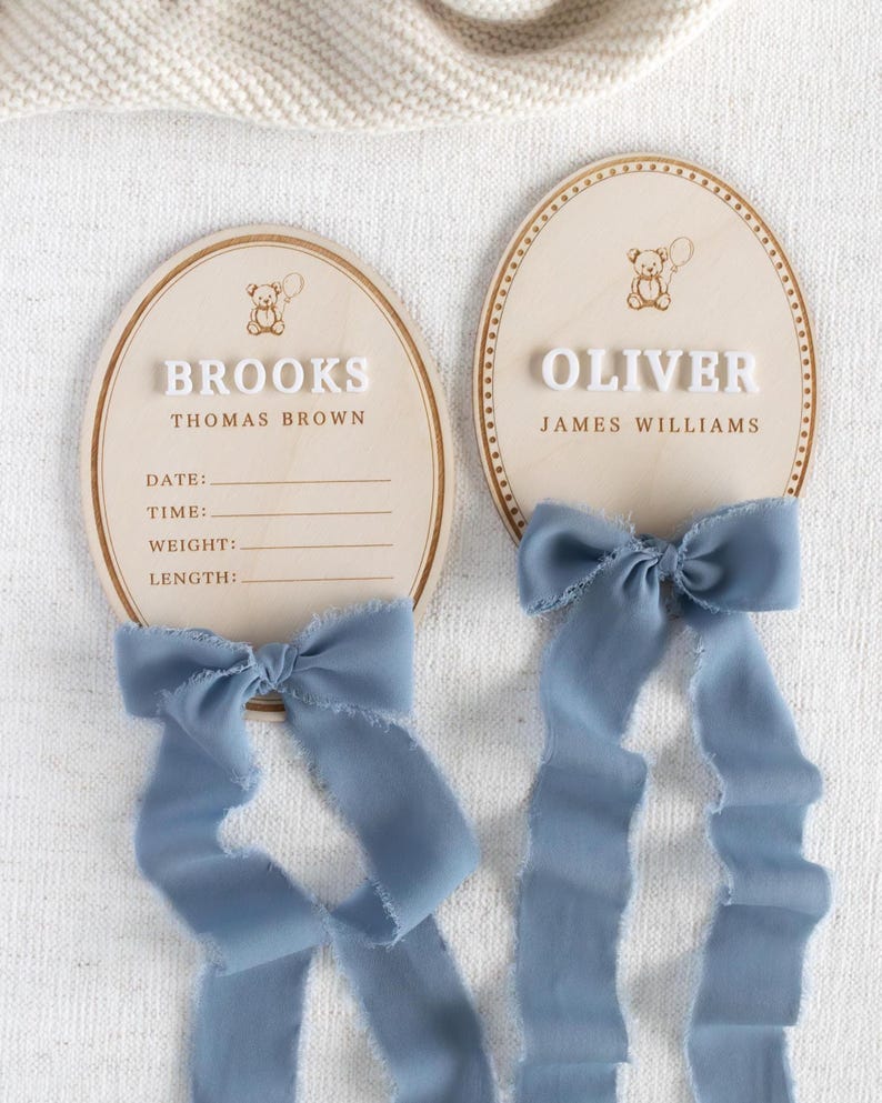 May include: Two wooden oval baby announcement plaques. One reads "BROOKS" and the other "OLIVER" in white block letters. Each has a teddy bear graphic and a light blue ribbon. The "BROOKS" plaque has lines for date, time, weight, and length.