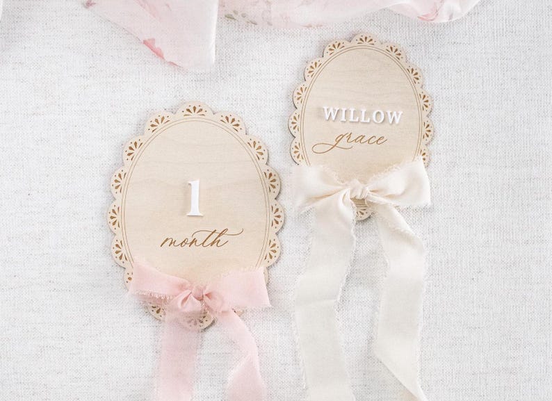 Lace Frame Milestone Marker with Scallop Border. Coquette Bow Detail with block font first name and script middle name or last name or both. Each Lace Frame is adorned with a silk chiffon ribbon in the color light pink or ivory. White acrylic milestone markers with the monthly number interchangeable in acrylic. Matching lace Frame name plate that can be used as a newborn hospital announcement. Milestone marker for girl, bow milestone card for girl, hospital name announcement for girl, wooden