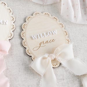 Lace Frame Milestone Marker with Scallop Border. Coquette Bow Detail with block font first name and script middle name or last name or both. Each Lace Frame is adorned with a silk chiffon ribbon in the color light pink or ivory. White acrylic milestone markers with the monthly number interchangeable in acrylic. Matching lace Frame name plate that can be used as a newborn hospital announcement. Milestone marker for girl, bow milestone card for girl, hospital name announcement for girl, wooden