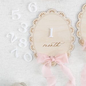 Lace Frame Milestone Marker with Scallop Border. Coquette Bow Detail with block font first name and script middle name or last name or both. Each Lace Frame is adorned with a silk chiffon ribbon in the color light pink or ivory. White acrylic milestone markers with the monthly number interchangeable in acrylic. Matching lace Frame name plate that can be used as a newborn hospital announcement. Milestone marker for girl, bow milestone card for girl, hospital name announcement for girl, wooden
