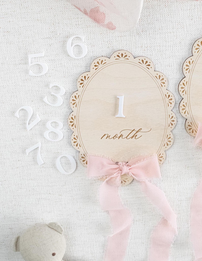 Lace Frame Milestone Marker with Scallop Border. Coquette Bow Detail with block font first name and script middle name or last name or both. Each Lace Frame is adorned with a silk chiffon ribbon in the color light pink or ivory. White acrylic milestone markers with the monthly number interchangeable in acrylic. Matching lace Frame name plate that can be used as a newborn hospital announcement. Milestone marker for girl, bow milestone card for girl, hospital name announcement for girl, wooden