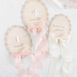 Lace Frame Milestone Marker with Scallop Border. Coquette Bow Detail with block font first name and script middle name or last name or both. Each Lace Frame is adorned with a silk chiffon ribbon in the color light pink or ivory. White acrylic milestone markers with the monthly number interchangeable in acrylic. Matching lace Frame name plate that can be used as a newborn hospital announcement. Milestone marker for girl, bow milestone card for girl, hospital name announcement for girl, wooden