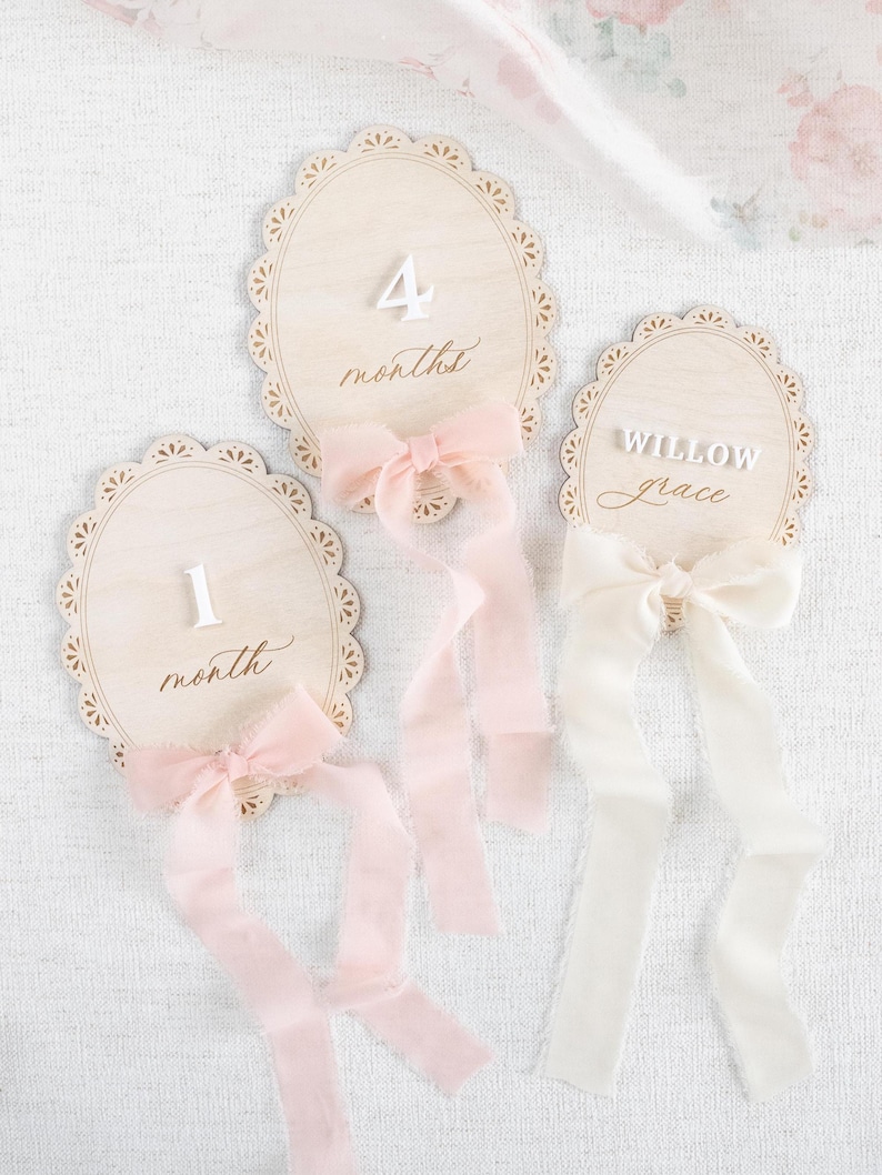 Lace Frame Milestone Marker with Scallop Border. Coquette Bow Detail with block font first name and script middle name or last name or both. Each Lace Frame is adorned with a silk chiffon ribbon in the color light pink or ivory. White acrylic milestone markers with the monthly number interchangeable in acrylic. Matching lace Frame name plate that can be used as a newborn hospital announcement. Milestone marker for girl, bow milestone card for girl, hospital name announcement for girl, wooden