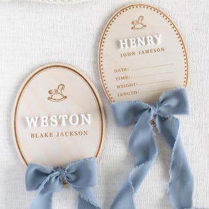 May include: Two wooden oval baby announcement plaques. Each plaque has a light wood grain finish, a decorative border, and a rocking horse illustration. One plaque reads "WESTON" and "BLAKE JACKSON", the other reads "HENRY" and "JOHN JAMESON". Both have blue ribbon bows.
