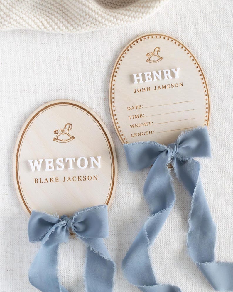 May include: Two wooden oval baby announcement plaques. Each plaque has a light wood grain finish, a decorative border, and a rocking horse illustration. One plaque reads "WESTON" and "BLAKE JACKSON", the other reads "HENRY" and "JOHN JAMESON". Both have blue ribbon bows.