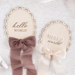 May include: Two wooden oval signs with scalloped edges and the text "hello world" in different fonts and styles. One sign has a brown ribbon tied around it, and the other has a white ribbon.