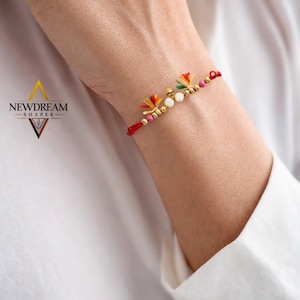 Peut inclure: Un bracelet en fil rouge orné de deux petites breloques en forme de fleurs orange et vertes, deux perles sphériques blanches et plusieurs petites perles dorées métalliques. Le bracelet est noué et porte le texte NEWDREAM SHAPER.
