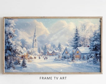 Op de afbeelding: Een ingelijst winterlandschap schilderij van een besneeuwd dorp met een kerktoren, huizen en bomen. Het schilderij heeft een lichtblauwe lucht met vallende witte sneeuw. De lijst is van licht hout. De tekst "FRAME TV ART" staat onder het schilderij.