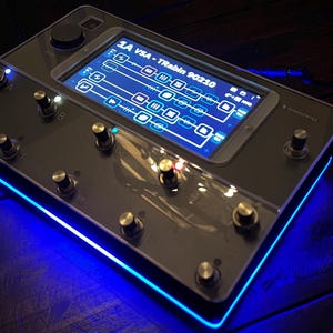 Peut inclure: Un appareil électronique noir avec un écran affichant une interface utilisateur. L'appareil comporte plusieurs boutons et molettes, avec une bande lumineuse LED bleue le long du bord inférieur. L'écran affiche le texte "1A V54 - Trabin 90210".