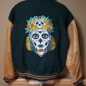 May include: A dark green varsity jacket with brown leather sleeves. The back of the jacket features an embroidered design of a woman's face with a skull and flowers.