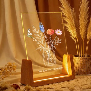 May include: A wooden-based LED night light. The clear acrylic panel displays a floral arrangement in a jar, with names and the text "We love you Mummy x". The light provides a warm, inviting glow.