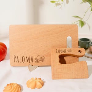 Puede incluir: Una tabla de cortar rectangular de madera y una herramienta de corte a juego, ambas con el nombre PALOMA grabado y un pequeño gráfico de sándwich. Los artículos de madera clara se muestran junto a un tomate rojo y dos galletas.