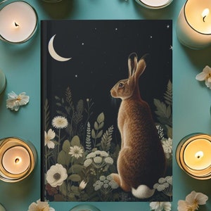 May include: A black journal with a brown rabbit looking up at a crescent moon and stars on a dark blue background. The rabbit is surrounded by white flowers and green foliage.