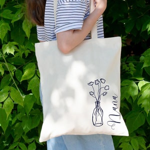Pu&ograve; includere: Una borsa tote in tela bianca con un disegno a linea nera di fiori in un vaso e la parola "Mama" scritta verticalmente sul lato destro.