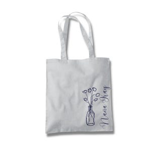 Pu&ograve; includere: Una borsa tote bianca con un disegno a linea blu di un vaso con fiori e il testo "Nana Ray" sul lato.