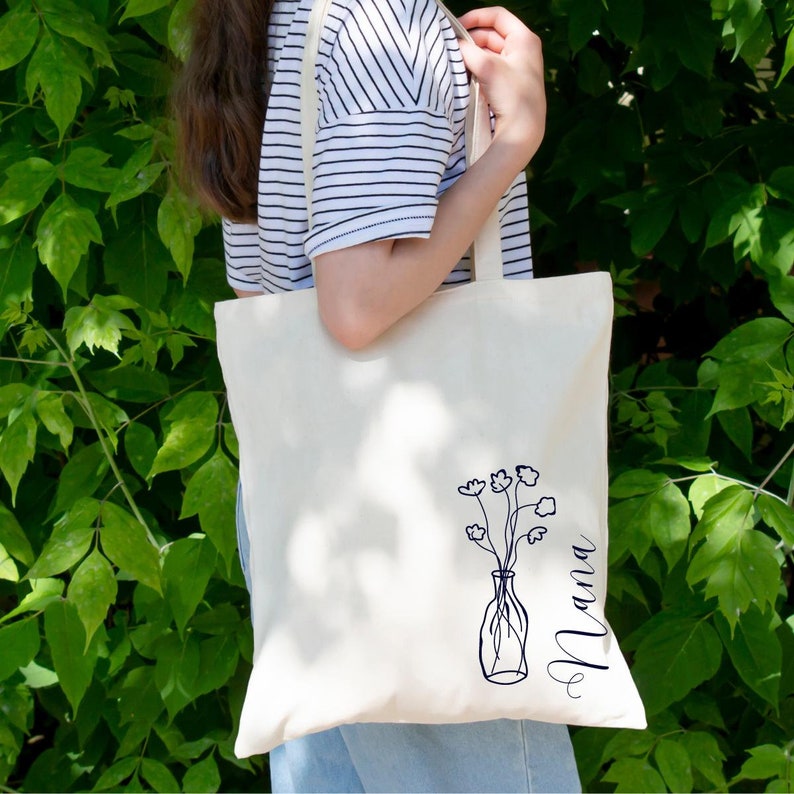 Pu&ograve; includere: Una borsa tote in tela bianca con un disegno a linea nera di fiori in un vaso e la parola "Mama" scritta verticalmente sul lato destro.