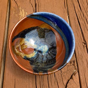 May include: A handmade ceramic bowl with a unique, abstract design. The bowl features a vibrant color palette, including shades of blue, brown, and black. The interior has a swirling pattern, and the rim is a deep blue. The bowl is set against a rustic wooden surface.