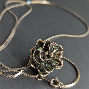May include: A silver necklace featuring a flower pendant with iridescent green and black enamel. The flower pendant is the focal point, with detailed petals. The chain is delicate, and the clasp is visible.