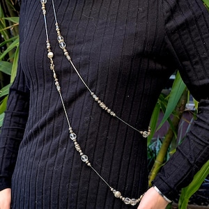 May include: A long, multi-strand necklace featuring clear faceted glass beads and metallic silver-toned beads arranged in a repeating pattern along thin, flexible wires, worn diagonally across a black ribbed long-sleeve dress.