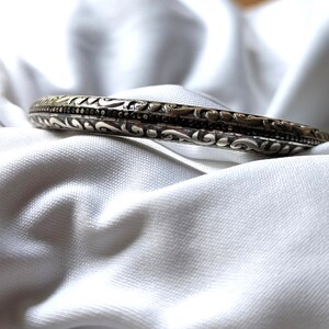 May include: A silver-toned bangle bracelet with intricate, swirling patterns and a dark band. The bracelet is set against a white, textured background, highlighting its detailed design and metallic sheen.
