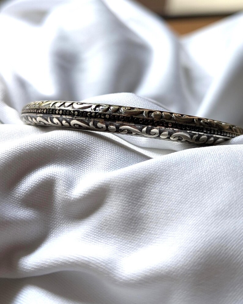 May include: A silver-toned bangle bracelet with intricate, swirling patterns and a dark band. The bracelet is set against a white, textured background, highlighting its detailed design and metallic sheen.