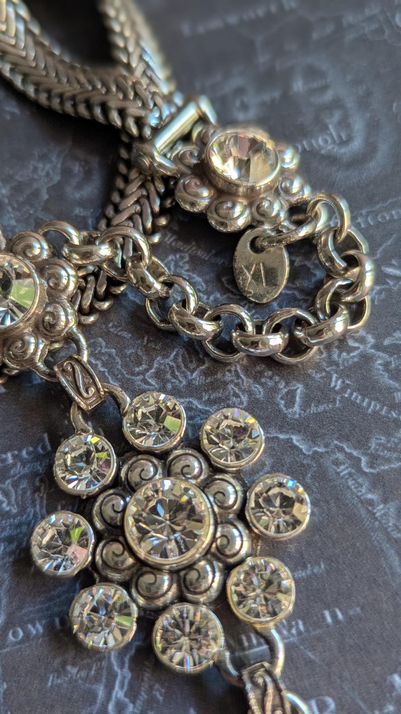 Peut inclure: Collier argent&eacute; avec un pendentif floral et des cristaux transparents &eacute;tincelants. Le collier pr&eacute;sente un motif de cha&icirc;ne et une petite &eacute;tiquette. Le pendentif a un grand cristal central entour&eacute; de petits cristaux.