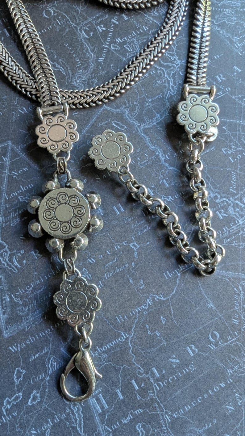 Peut inclure: Collier argent&eacute; avec un motif floral. Le collier est compos&eacute; d'une cha&icirc;ne avec un fermoir en forme de fleur et de pendentifs en forme de fleur. Le fond est un motif de carte.