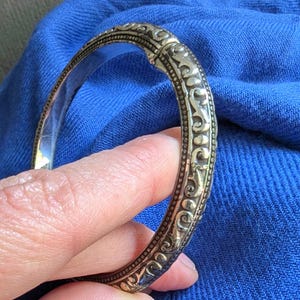 May include: Ornate gold-toned bangle bracelet with detailed scrollwork and a hinged clasp. The bracelet's design features a series of repeating patterns and a polished interior. The bracelet is held against a blue fabric background.