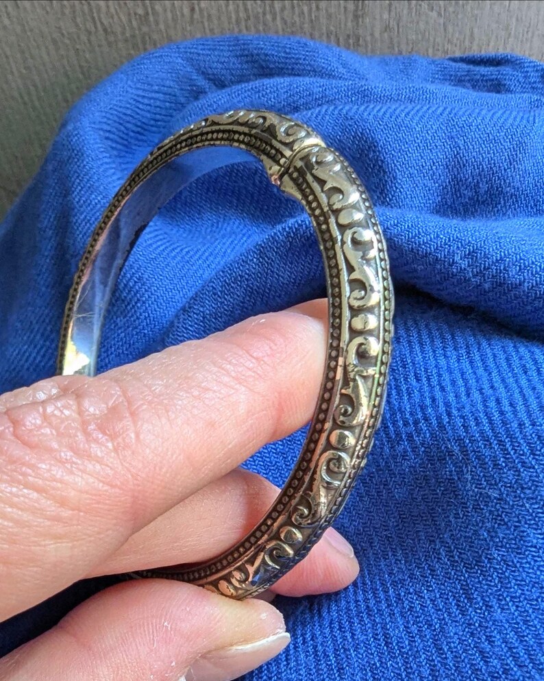 May include: Ornate gold-toned bangle bracelet with detailed scrollwork and a hinged clasp. The bracelet's design features a series of repeating patterns and a polished interior. The bracelet is held against a blue fabric background.